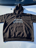 Ein schwarzer Kapuzenpullover Save The Classics von SaveTheClassics mit der Aufschrift "Save the Classics" in weißen gotischen Buchstaben ist auf der Motorhaube eines weißen Autos abgebildet, wobei derselbe Satz auf der Windschutzscheibe dahinter zu sehen ist.