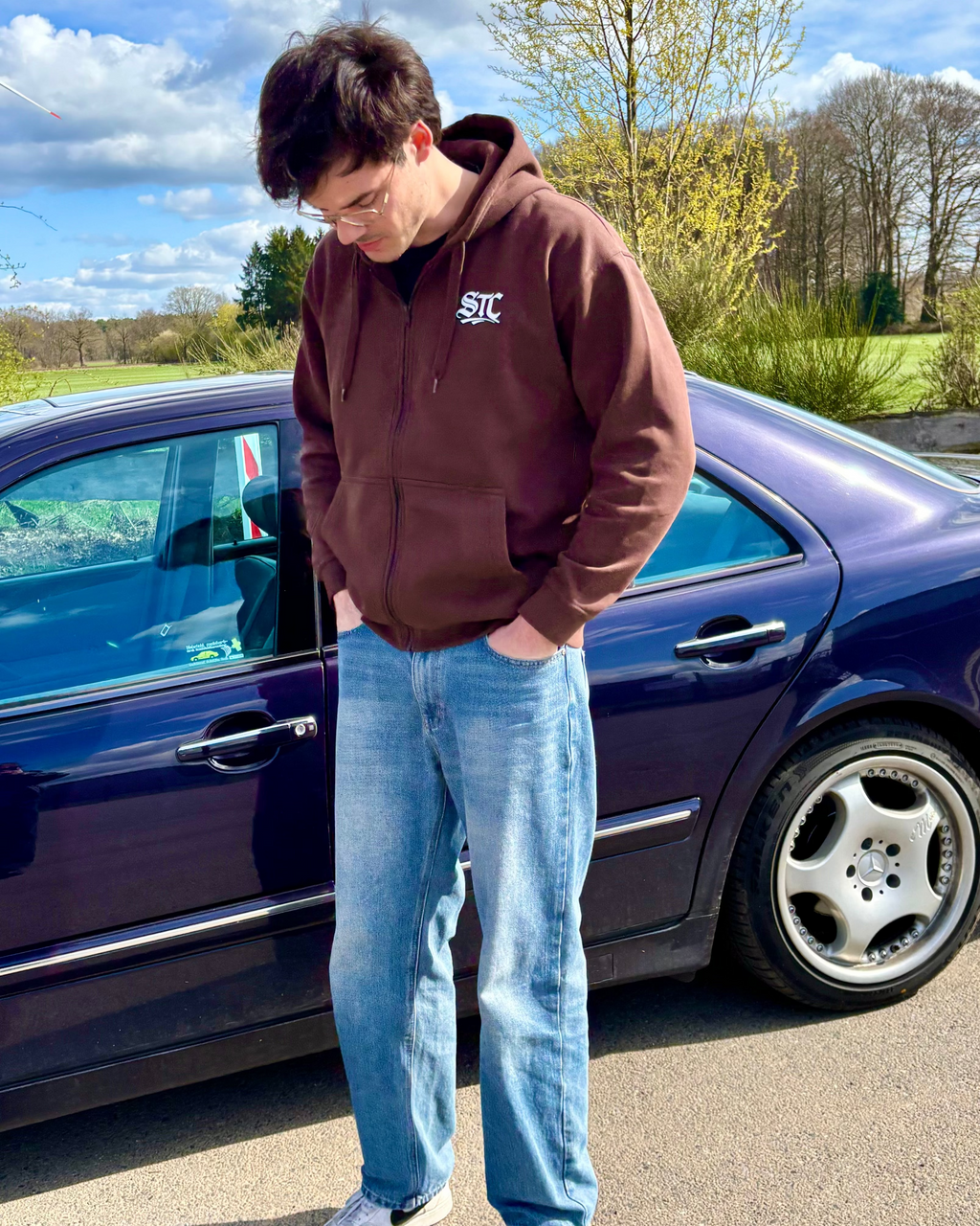 Ein brauner SaveTheClassics Zip Hoodie mit kleinem SIC-Logo auf der linken Brust liegt auf der dunkelblauen Motorhaube eines Autos und unterstreicht die automobile Streetwear mit der Windschutzscheibe und dem Innenraum im Hintergrund.