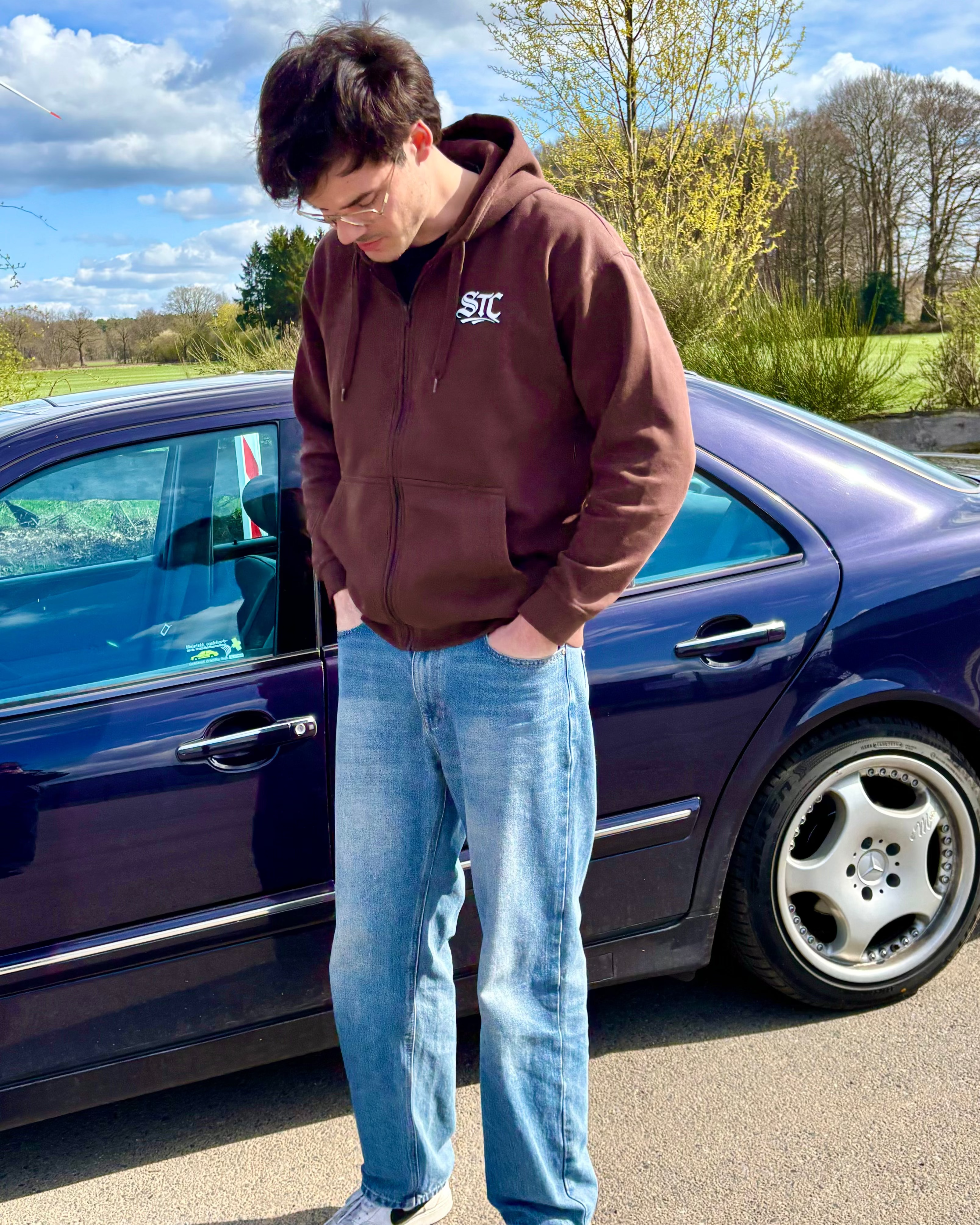 Ein brauner SaveTheClassics Zip Hoodie mit kleinem SIC-Logo auf der linken Brust liegt auf der dunkelblauen Motorhaube eines Autos und unterstreicht die automobile Streetwear mit der Windschutzscheibe und dem Innenraum im Hintergrund.