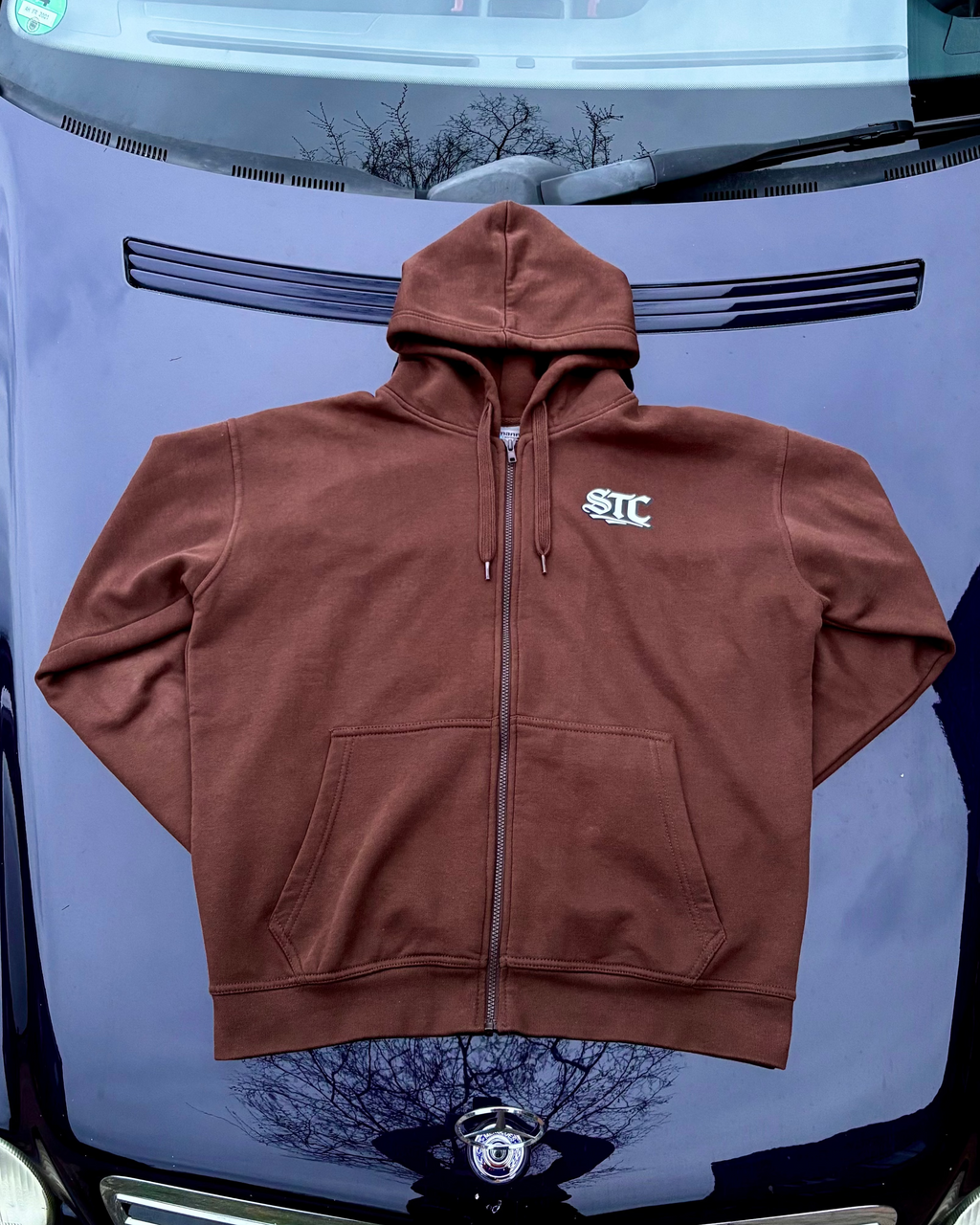 Ein brauner SaveTheClassics Zip Hoodie mit kleinem SIC-Logo auf der linken Brust liegt auf der dunkelblauen Motorhaube eines Autos und unterstreicht die automobile Streetwear mit der Windschutzscheibe und dem Innenraum im Hintergrund.