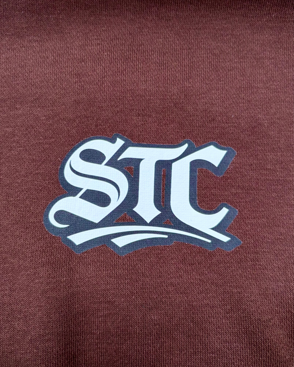 Ein brauner SaveTheClassics Zip Hoodie mit kleinem SIC-Logo auf der linken Brust liegt auf der dunkelblauen Motorhaube eines Autos und unterstreicht die automobile Streetwear mit der Windschutzscheibe und dem Innenraum im Hintergrund.