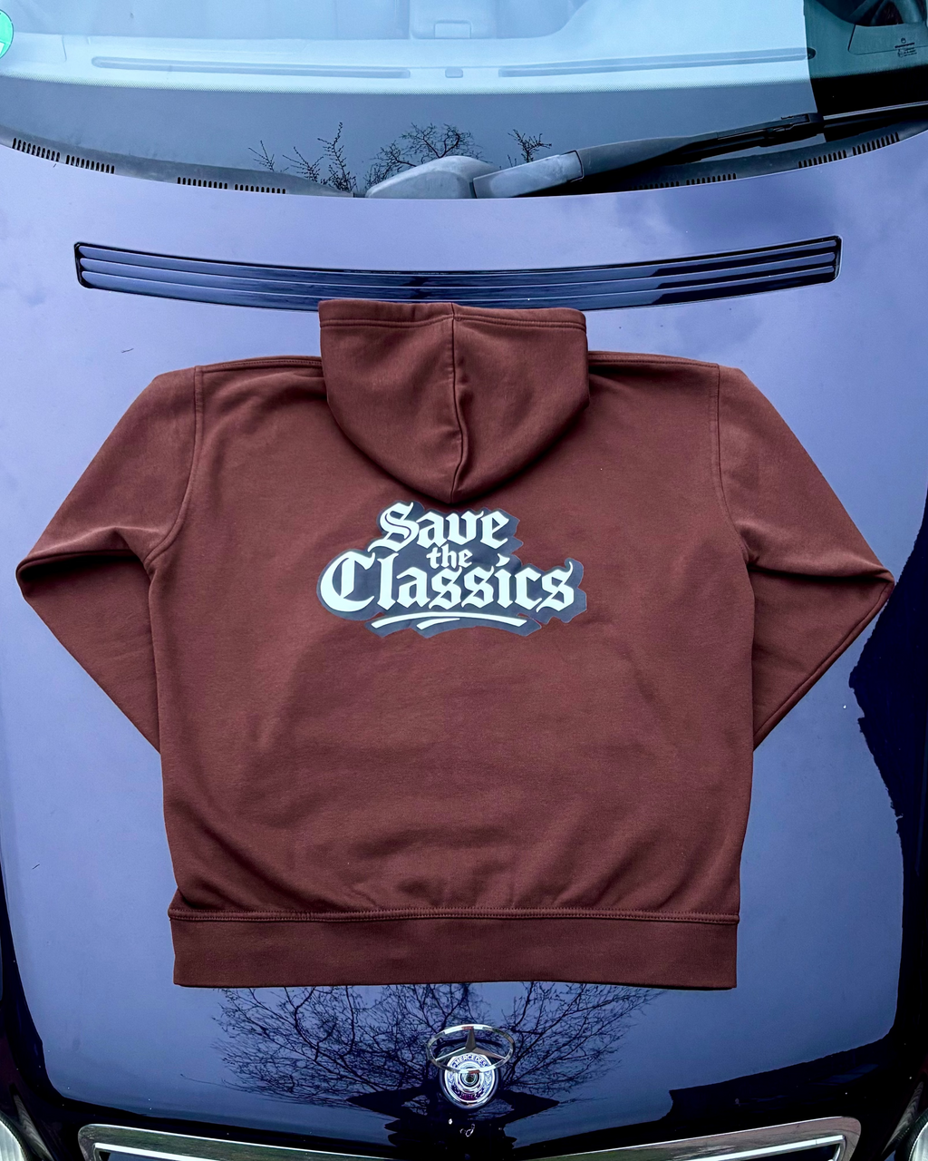 Ein brauner SaveTheClassics Zip Hoodie mit kleinem SIC-Logo auf der linken Brust liegt auf der dunkelblauen Motorhaube eines Autos und unterstreicht die automobile Streetwear mit der Windschutzscheibe und dem Innenraum im Hintergrund.