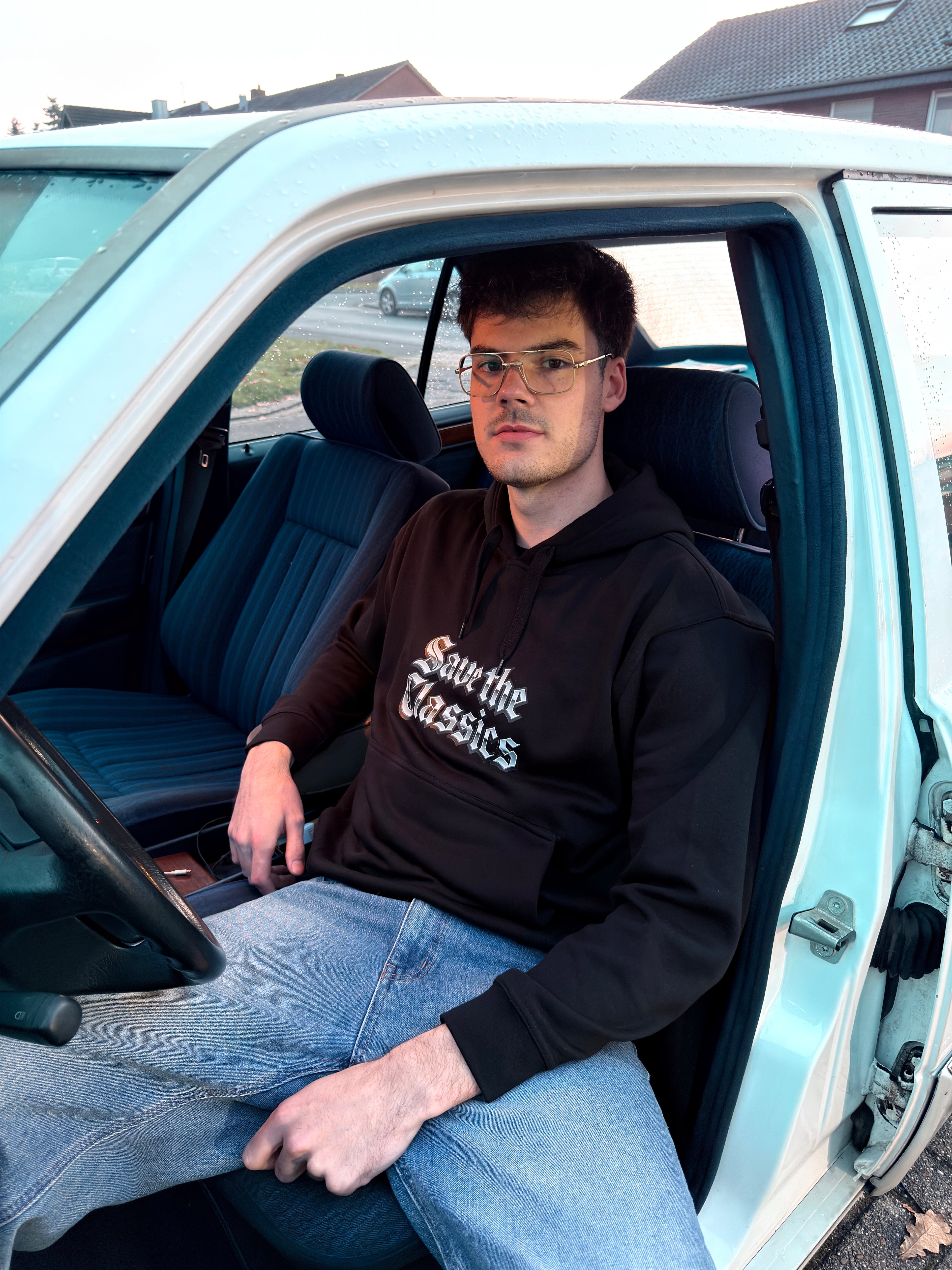 Ein schwarzer Kapuzenpullover Save The Classics von SaveTheClassics mit der Aufschrift "Save the Classics" in weißen gotischen Buchstaben ist auf der Motorhaube eines weißen Autos abgebildet, wobei derselbe Satz auf der Windschutzscheibe dahinter zu sehen ist.