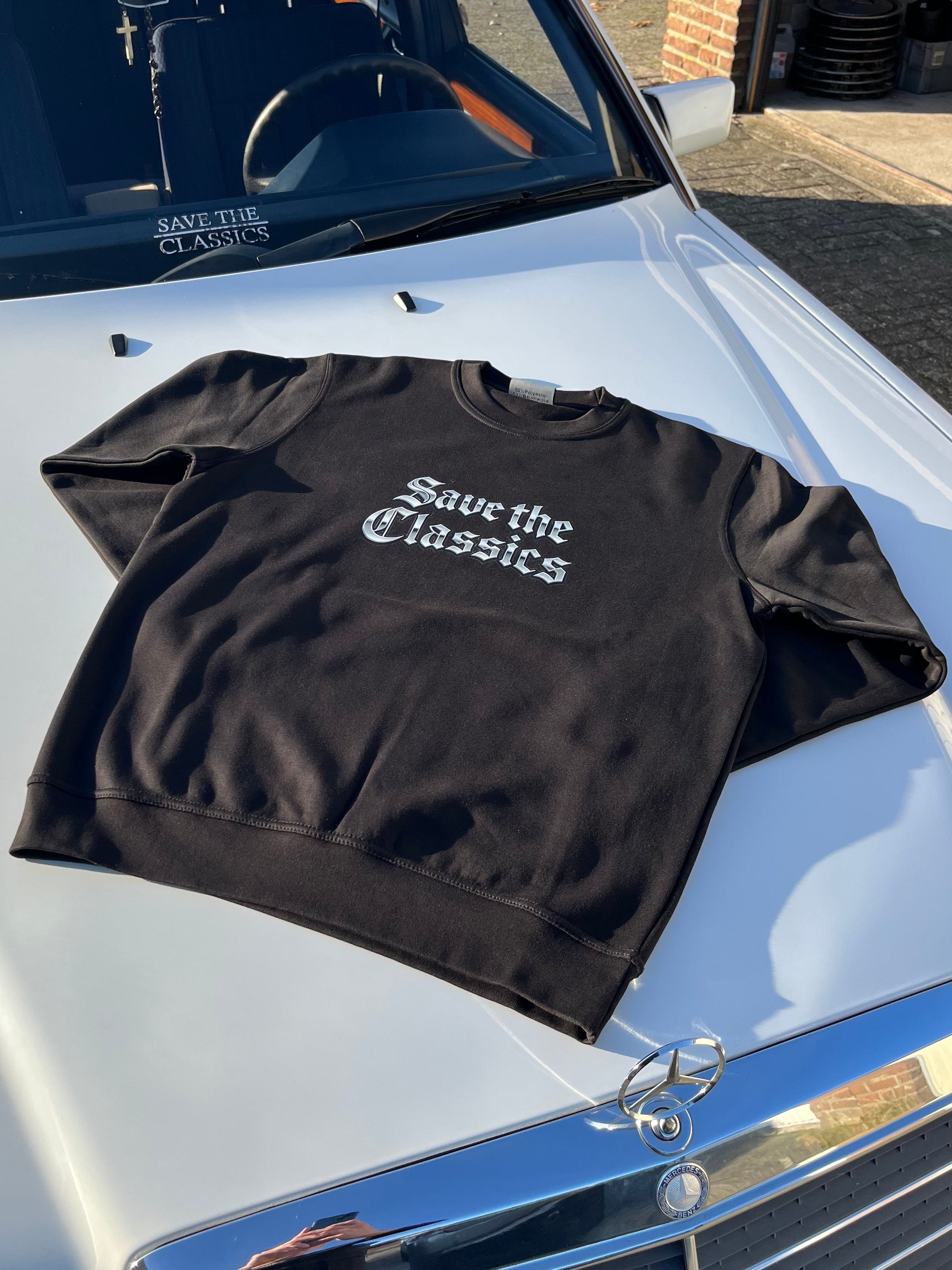 Das Sweatshirt Save the Classics von SaveTheClassics trägt einen auffälligen weißen Gothic-Schriftzug und wird auf einem weißen Mercedes-Benz abgebildet, der unter dramatischem Sonnenlicht und Schatten Streetwear mit automobiler Kultur verschmilzt.